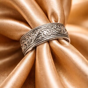 Può includere: Un anello d'argento con un intricato motivo floreale. L'anello è posto su uno sfondo di tessuto di raso dorato, creando un'estetica lussuosa ed elegante. Il design dell'anello presenta incisioni dettagliate.