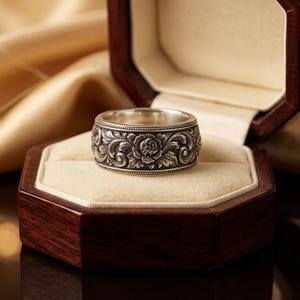 Puede incluir: Un anillo de plata con un intrincado diseño floral, con un motivo de rosa prominente. El anillo se muestra en una caja de joyería de madera oscura con un interior color crema, sobre un fondo de tela dorada.