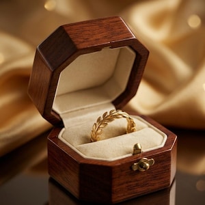 Può includere: Un anello d'oro con un design a foglie è contenuto in un portagioie ottagonale in legno aperto. L'interno del portagioie è beige. L'anello è il punto focale, con la scatola e lo sfondo sfocati.