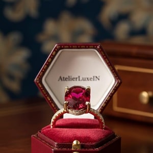 May include: A gold ring with a large, round, red gemstone, set in a red velvet jewelry box. The ring has a diamond-studded band. The jewelry box is red with gold accents and the text "AtelierLuxeIN".
