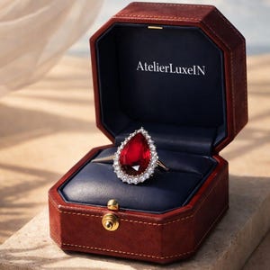 Op de afbeelding: Een gouden ring met een grote, peervormige, rode edelsteen omringd door witte stenen, gepresenteerd in een open, bruine leren sieradendoos met een blauw interieur. De tekst "AtelierLuxeIN" is zichtbaar.