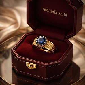 Puede incluir: Un anillo de oro con una piedra preciosa grande, redonda y azul oscuro, engastada en una banda ornamentada y grabada. El anillo se muestra en un joyero burdeos abierto con un cierre dorado. El texto "AtelierLuxeIN" está en el interior.