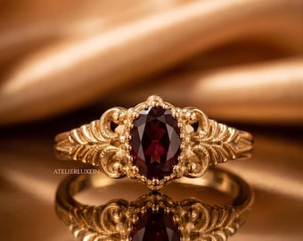 May include: A gold ring with an oval, deep red gemstone. The ring has an ornate, filigree design around the gemstone and on the band. The ring is reflected in a shiny surface. The text "ATELIERLUXEIN" is visible.