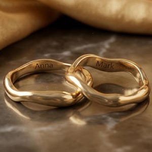 May include: Two gold-colored rings interlocked, with a wavy design. One ring is engraved with the name "Anna", and the other with "Mark". The rings are displayed on a reflective surface.