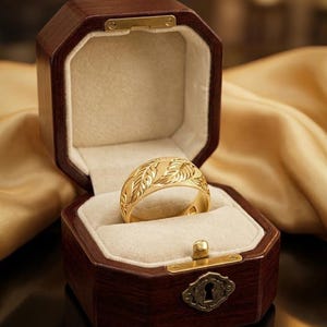 May include: A gold ring with a leaf pattern sits inside a dark brown, octagonal jewelry box with a cream-colored interior. The ring is the focal point, showcasing intricate details. The box is open, revealing the ring against a soft, neutral backdrop.