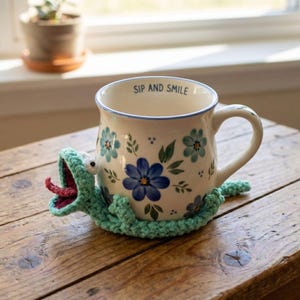 Może przedstawiać: Ceramiczny kubek z białym tłem, niebieskimi wzorami kwiatowymi i napisem SIP AND SMILE na wewnętrznej krawędzi. Kubek jest otoczony jasną, szydełkowaną osłoną w kształcie stworzenia z otwartym pyszczkiem, różowym językiem i widocznymi oczami.