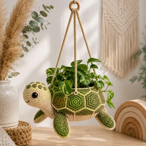 Puede incluir: Una maceta colgante de ganchillo con forma de tortuga, cabeza beige claro y caparazón con patrón hexagonal verde. La maceta está suspendida por tres cordones trenzados unidos a un anillo de madera y contiene una planta verde de hojas variegadas.