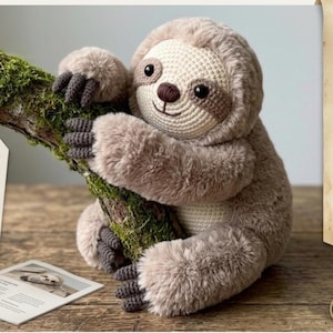 Puede incluir: Un peluche de perezoso marrón claro con cara y cuerpo de ganchillo, abrazando una rama cubierta de musgo. El perezoso tiene ojos oscuros y una expresión sonriente. El juguete mide aproximadamente 30 cm de alto.