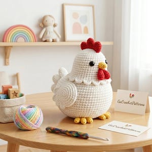 May include: A large, white crocheted chicken with a red comb and wattle, black eyes, and yellow feet sits on a wooden table. A ball of rainbow yarn and a crochet hook are nearby. The scene includes a shelf with a rainbow and a doll.