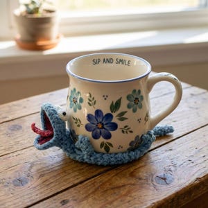May include: A cream-colored ceramic mug featuring blue floral patterns and the text SIP AND SMILE printed on the inner rim. The mug is wrapped in a blue crocheted creature cozy with a wide-open mouth, a pink tongue, and two white eyes.