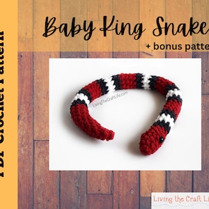 Può includere: Un serpente reale bambino all'uncinetto con strisce rosse, nere e bianche. Il serpente è arrotolato in un cerchio. L'immagine include il testo "Baby King Snake + bonus pattern".