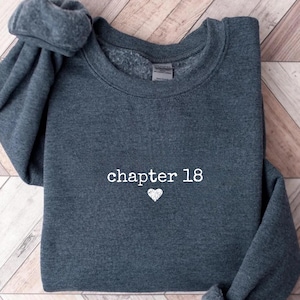Peut inclure: Sweat-shirt gris foncé avec l'inscription "chapter 18" en blanc et un petit cœur blanc. Le sweat-shirt est plié sur une surface en bois.