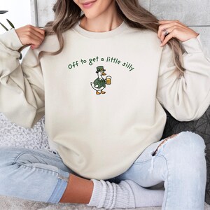 May include: Cream-colored sweatshirt with the embroidered phrase "off to get a little silly." The design features a cartoon duck wearing a green jacket and hat, holding a mug of beer. The sweatshirt is a casual, comfortable style.