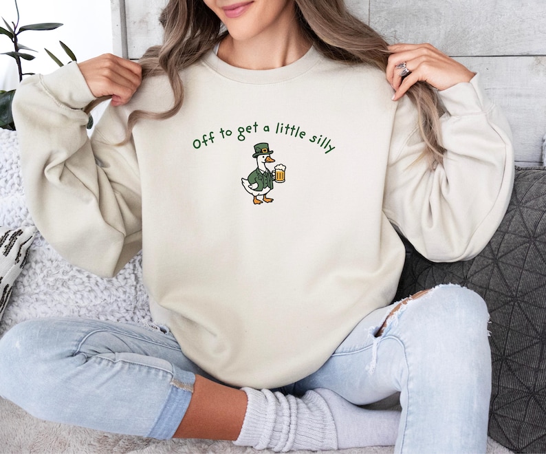 May include: Cream-colored sweatshirt with the embroidered phrase "off to get a little silly." The design features a cartoon duck wearing a green jacket and hat, holding a mug of beer. The sweatshirt is a casual, comfortable style.