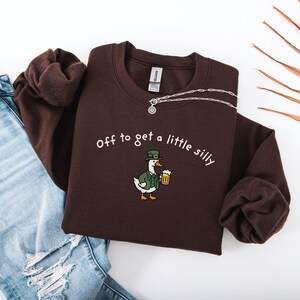 May include: A dark brown sweatshirt with the phrase "off to get a little silly" in white script. The sweatshirt features an embroidered cartoon duck wearing a green hat and jacket, holding a beer. A silver necklace is draped across the top.