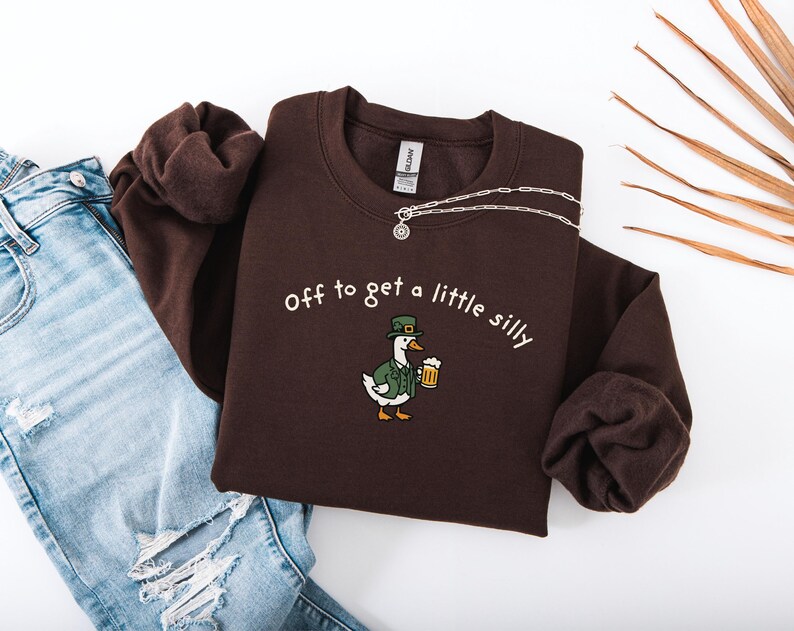 May include: A dark brown sweatshirt with the phrase "off to get a little silly" in white script. The sweatshirt features an embroidered cartoon duck wearing a green hat and jacket, holding a beer. A silver necklace is draped across the top.