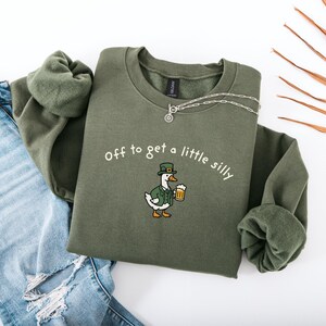 May include: Olive green sweatshirt with the phrase "off to get a little silly" and a cartoon duck wearing a green hat and jacket, holding a beer mug. The sweatshirt is paired with blue jeans and a silver necklace.