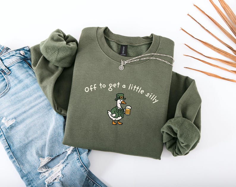 May include: Olive green sweatshirt with the phrase "off to get a little silly" and a cartoon duck wearing a green hat and jacket, holding a beer mug. The sweatshirt is paired with blue jeans and a silver necklace.