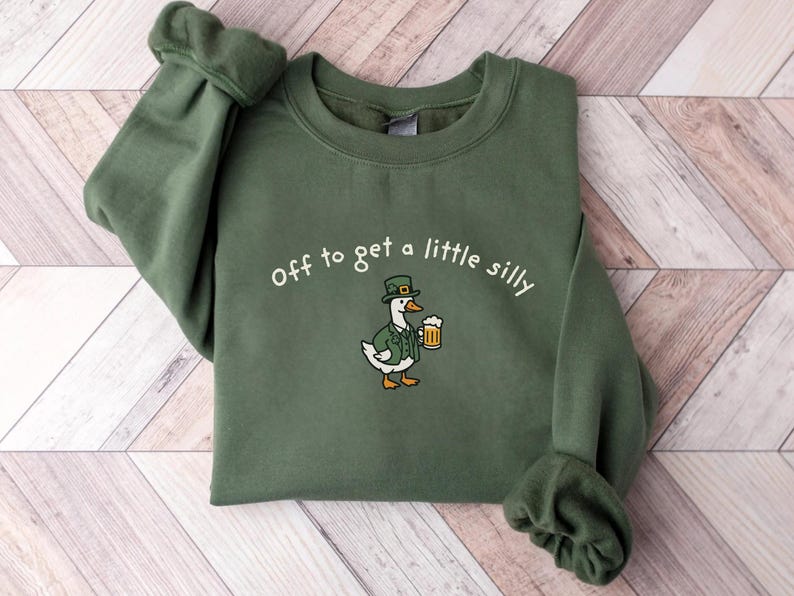 May include: Green sweatshirt with the embroidered phrase "Off to get a little silly." The design features a cartoon goose wearing a green hat and jacket, holding a mug of beer. The sweatshirt is a solid green color.