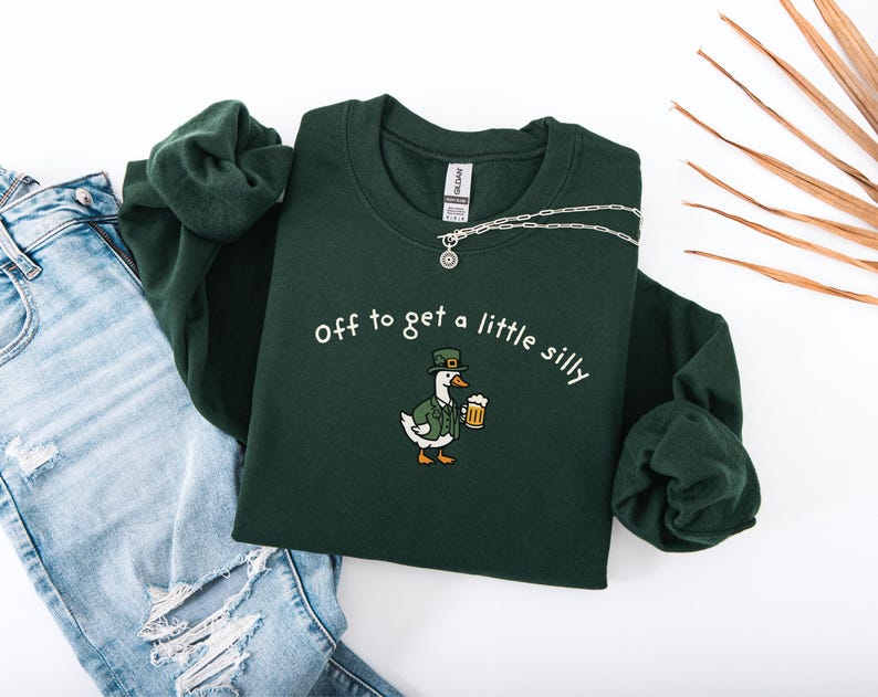 May include: A dark green sweatshirt with the phrase "Off to get a little silly" and a cartoon goose wearing a hat and holding a beer. A silver necklace is draped over the sweatshirt, which is paired with blue jeans.