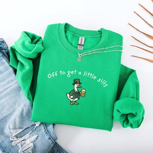 May include: A green sweatshirt with the phrase "off to get a little silly" embroidered in white. The sweatshirt features a cartoon duck wearing a leprechaun hat and holding a beer. A silver necklace is draped on the sweatshirt. Blue jeans are next to the sweatshirt.