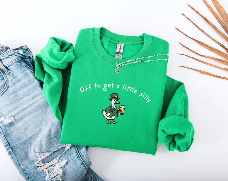 May include: A green sweatshirt with the phrase "off to get a little silly" embroidered in white. The sweatshirt features a cartoon duck wearing a leprechaun hat and holding a beer. A silver necklace is draped on the sweatshirt. Blue jeans are next to the sweatshirt.