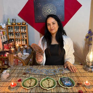 May include: A person sits at a bamboo mat table with three leaf-shaped ceramic dishes, one holding a tarot deck. A lit oil lamp, two small candles, and a wooden shelf filled with small glass bottles are present. The person holds a circular fan.