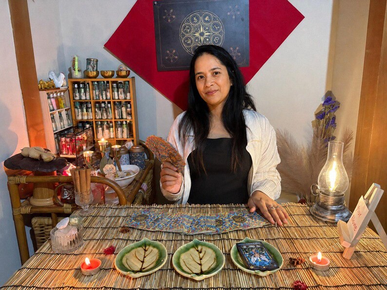 May include: A person sits at a bamboo mat table with three leaf-shaped ceramic dishes, one holding a tarot deck. A lit oil lamp, two small candles, and a wooden shelf filled with small glass bottles are present. The person holds a circular fan.
