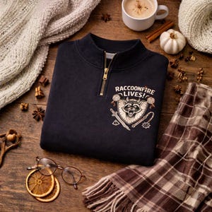 Könnte beinhalten: Marineblaues Sweatshirt mit goldenem Reißverschluss und dem Text "RACCOONFIRE LIVES!". Die Grafik zeigt einen Waschbären auf einem Schild mit Schwert und Flegel. Herbstliche Accessoires umgeben das Sweatshirt.