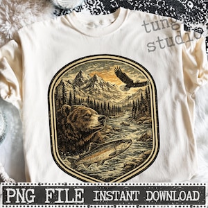 Puede incluir: Camiseta de color crema con un gráfico temático de la naturaleza. El diseño incluye un oso, un pez, un águila, un río y montañas dentro de un marco circular. El texto "PNG FILE INSTANT DOWNLOAD" está en la parte inferior.