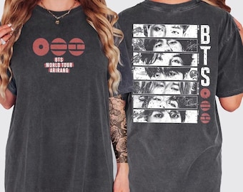 May include: Dark gray t-shirt featuring the text "BTS WORLD TOUR ARIRANG" in white and red. The front of the shirt has a circular design. The back of the shirt has a black and white image of faces and the letters "BTS".