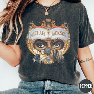 May include: Dark gray t-shirt featuring a graphic of Michael Jackson's eyes and face, with the name "Michael Jackson" in a banner above. The design includes gold and blue accents, and various other images.