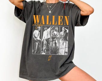 May include: Dark gray t-shirt featuring the name "WALLEN" in orange block letters above a black and white image of four men. The name "Morgan" is written in cursive below "WALLEN". The shirt has short sleeves and a relaxed fit.