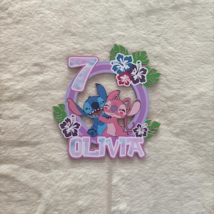 May include: A colorful cake topper featuring the characters Stitch and Angel, with the number 7 and the name Olivia. The design includes tropical flowers and leaves, with a purple and blue color scheme.