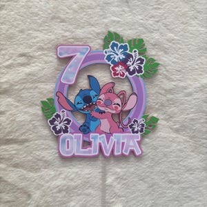 May include: A colorful cake topper featuring the characters Stitch and Angel embracing, with the number 7 and the name Olivia. The design includes a purple ring, tropical flowers, and green leaves, perfect for a birthday celebration.