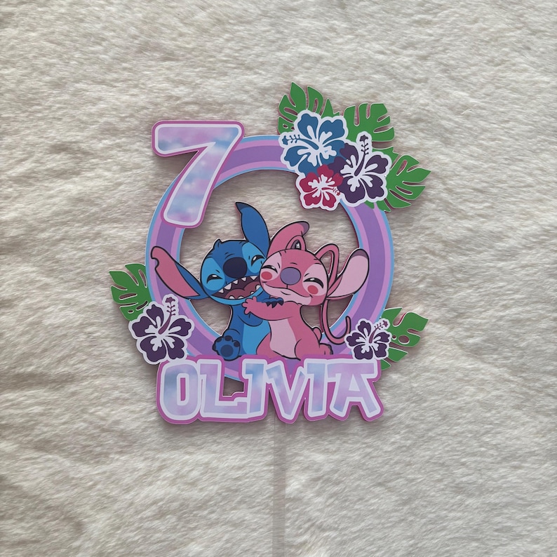 May include: A colorful cake topper featuring the characters Stitch and Angel embracing, with the number 7 and the name Olivia. The design includes a purple ring, tropical flowers, and green leaves, perfect for a birthday celebration.