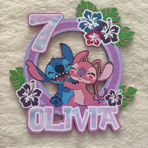 May include: A colorful birthday cake topper featuring the characters Stitch and Angel embracing. The number 7 is displayed within a circular frame, with the name "OLIVIA" below. Tropical flowers and leaves decorate the design.
