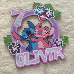 May include: A colorful cake topper featuring the characters Stitch and Angel from Lilo & Stitch. The design includes a number 7, the name "Olivia", tropical flowers, and a circular frame with a rainbow gradient.