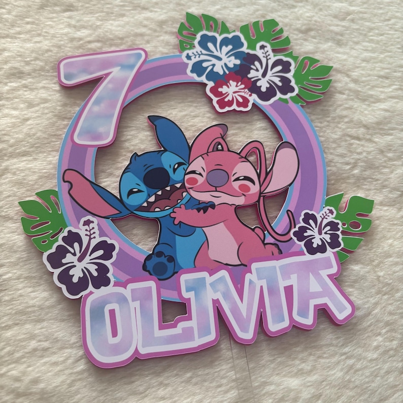 May include: A colorful cake topper featuring the characters Stitch and Angel from Lilo & Stitch. The design includes a number 7, the name "Olivia", tropical flowers, and a circular frame with a rainbow gradient.