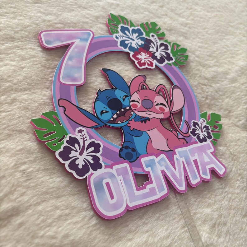 May include: A colorful cake topper featuring the characters Stitch and Angel from Lilo & Stitch. The design includes the number 7, the name "OLIVIA", and tropical flowers. The characters are hugging inside a circular frame.