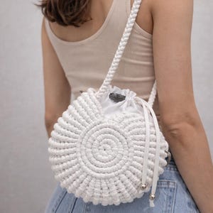 Puede incluir: Bolso blanco de macramé con correa trenzada. El bolso redondo presenta un diseño en espiral y un cierre de cordón. El bolso está hecho de un material grueso y tejido y tiene herrajes plateados.