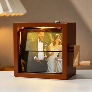 May include: A brown wooden photo cube with a hand crank, displaying a wedding photo. The cube has a light inside, illuminating the image of a smiling couple. The crank and handle are made of light-colored wood.