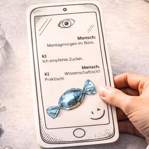 Könnte beinhalten: Eine rechteckige Karte mit einer schwarz-weißen Strichzeichnung einer Smartphone-Oberfläche, deutschem Text und einem einzelnen, in blaues Stanniolpapier eingewickelten Bonbon auf der Oberfläche. Der Text lautet: Mensch: Montagmorgen im Büro, KI: Ich empfehle Zucker, Mensch: Wissenschaftlich?, KI: Praktisch!