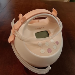 May include: A Spectra breast pump in white and light pink, featuring a handle and buttons labeled "Cycle", "Vacuum", and a power button. Clear tubing and a small digital display are also visible.