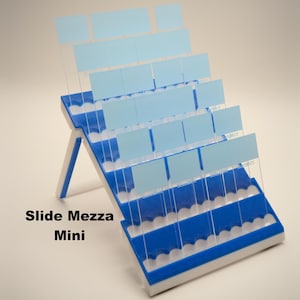 Può includere: Un Slide Mezza Mini blu e bianco, un organizer a più livelli con divisori trasparenti e inserti rettangolari azzurri. L'organizer ha una base bianca e un telaio blu, con il testo "Slide Mezza Mini" in nero.