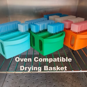 May include: Several colorful drying baskets are inside an oven. The baskets are light blue, green, and orange. Each basket holds multiple stacks of small, rectangular items in various colors. The text "Oven Compatible Drying Basket" is displayed.