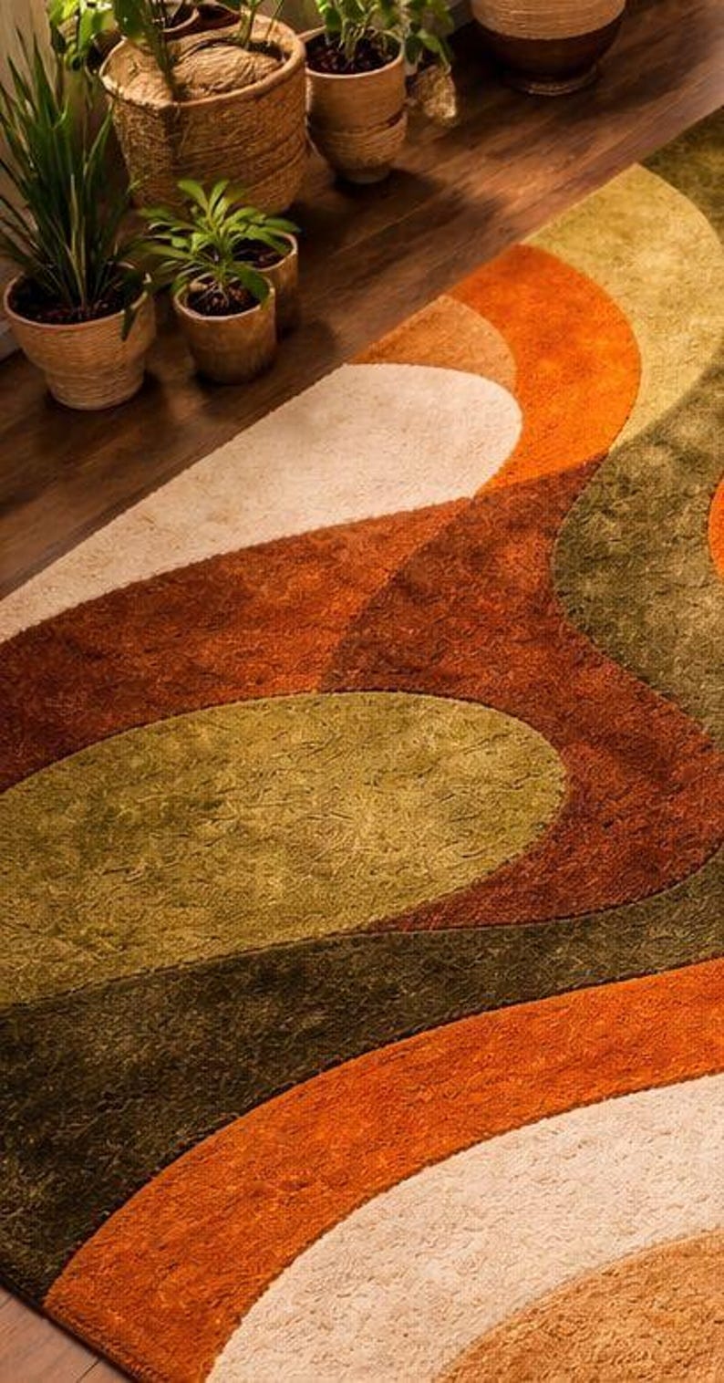 May include: A close-up of a retro-style rug with a geometric pattern. The rug features curved shapes in shades of orange, green, and cream. The rug is placed on a wooden floor, with several potted plants in the background.