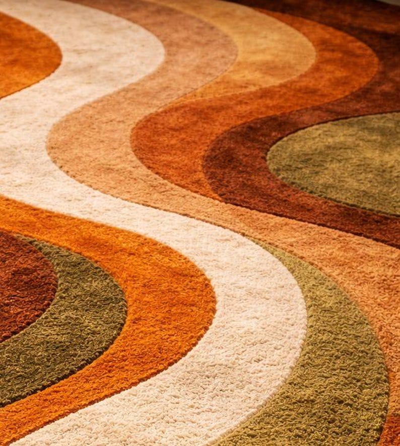 May include: Close-up of a retro-style rug with a wavy pattern. The rug features bands of color in shades of orange, brown, beige, and olive green. The texture appears plush and soft, with a vintage aesthetic.