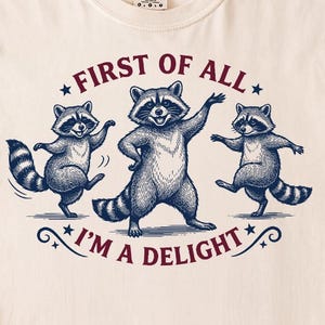 Könnte beinhalten: Cremefarbenes T-Shirt mit einer Grafik von drei tanzenden Waschbären. Der marineblaue Text lautet "FIRST OF ALL I'M A DELIGHT" mit Sternakzenten. Das Shirt ist aus weichem Material. Ein lustiges, lässiges Design.