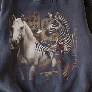 Puede incluir: Sudadera gris oscuro con una ilustración caprichosa de una cebra tatuando a un caballo. El caballo es blanco con rayas negras, y la cebra lleva un delantal. El fondo incluye diseños de tatuajes y una lámpara de escritorio.